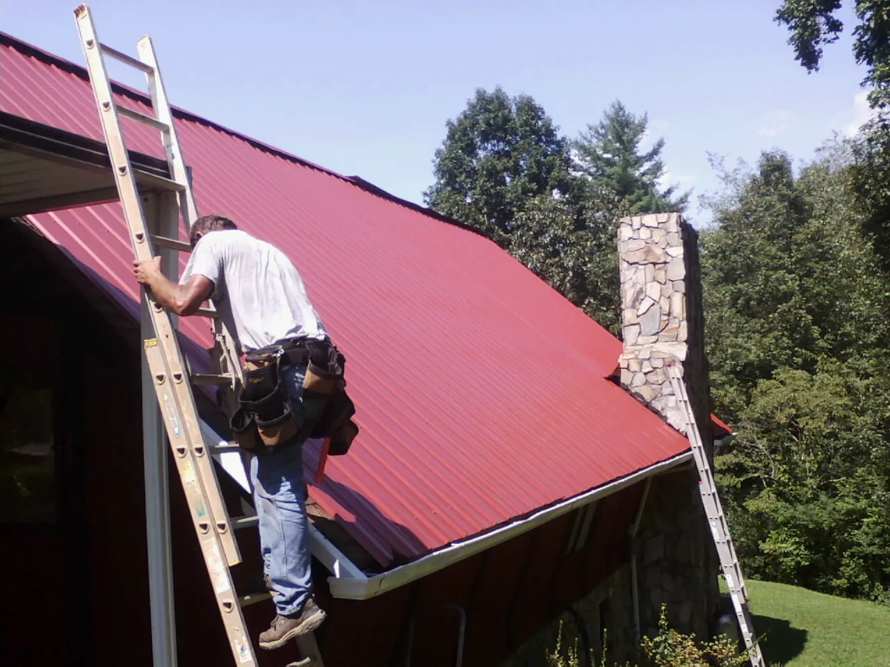 Roofer on a ladder beside a red metal roof and stone chimney — Soffit & Fascia Repair by Roofing Brothers in White Marsh